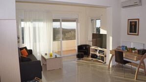Un salon moderne comprenant un canapé, une table basse et un coin repas avec un bureau et une chaise. Une grande fenêtre offre une vue sur l’extérieur.