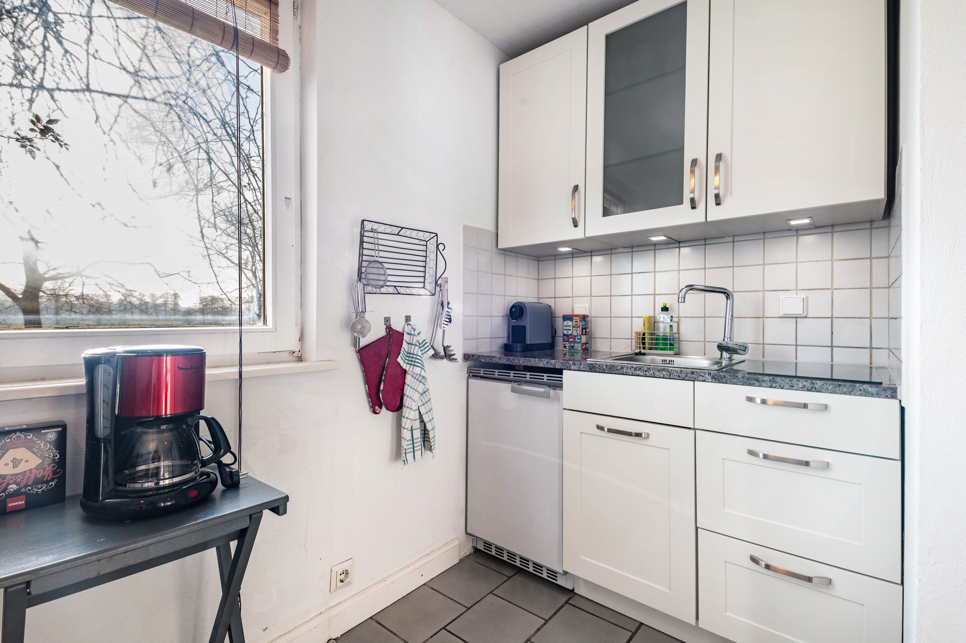 A kitchen with white cabinets, a coffee maker, a dishwasher, and a window with a view of trees.