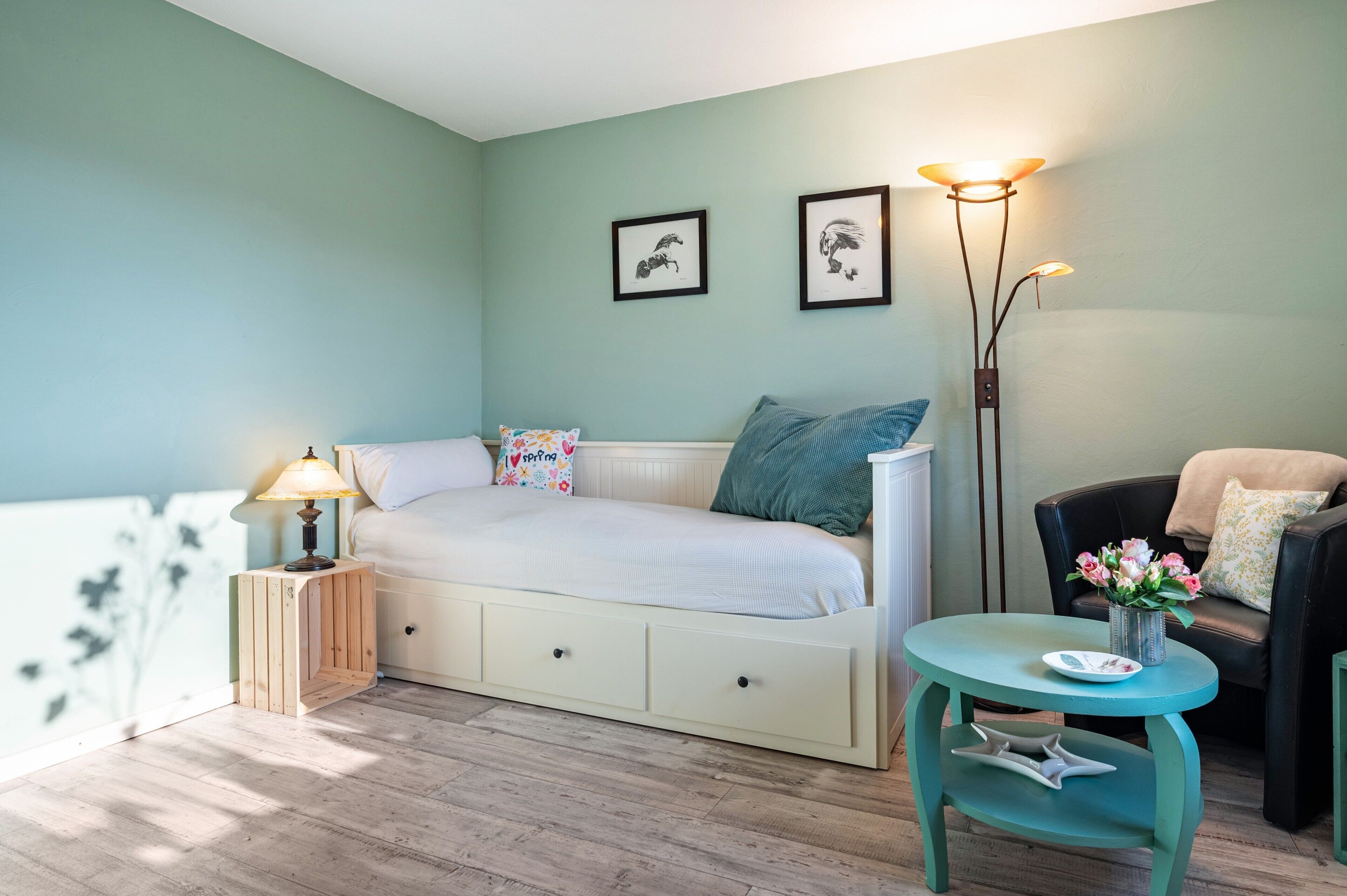 A cozy bedroom with a bed, bedside table, lamp, and a small table with a vase of flowers.