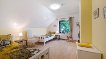 Quarto com duas camas, janela, radiador e uma mesinha.