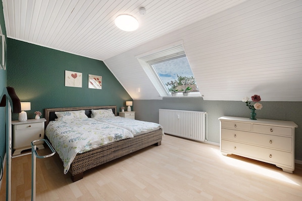 A bedroom with a sloped ceiling, a bed, a dresser, a nightstand, a window, and a radiator.