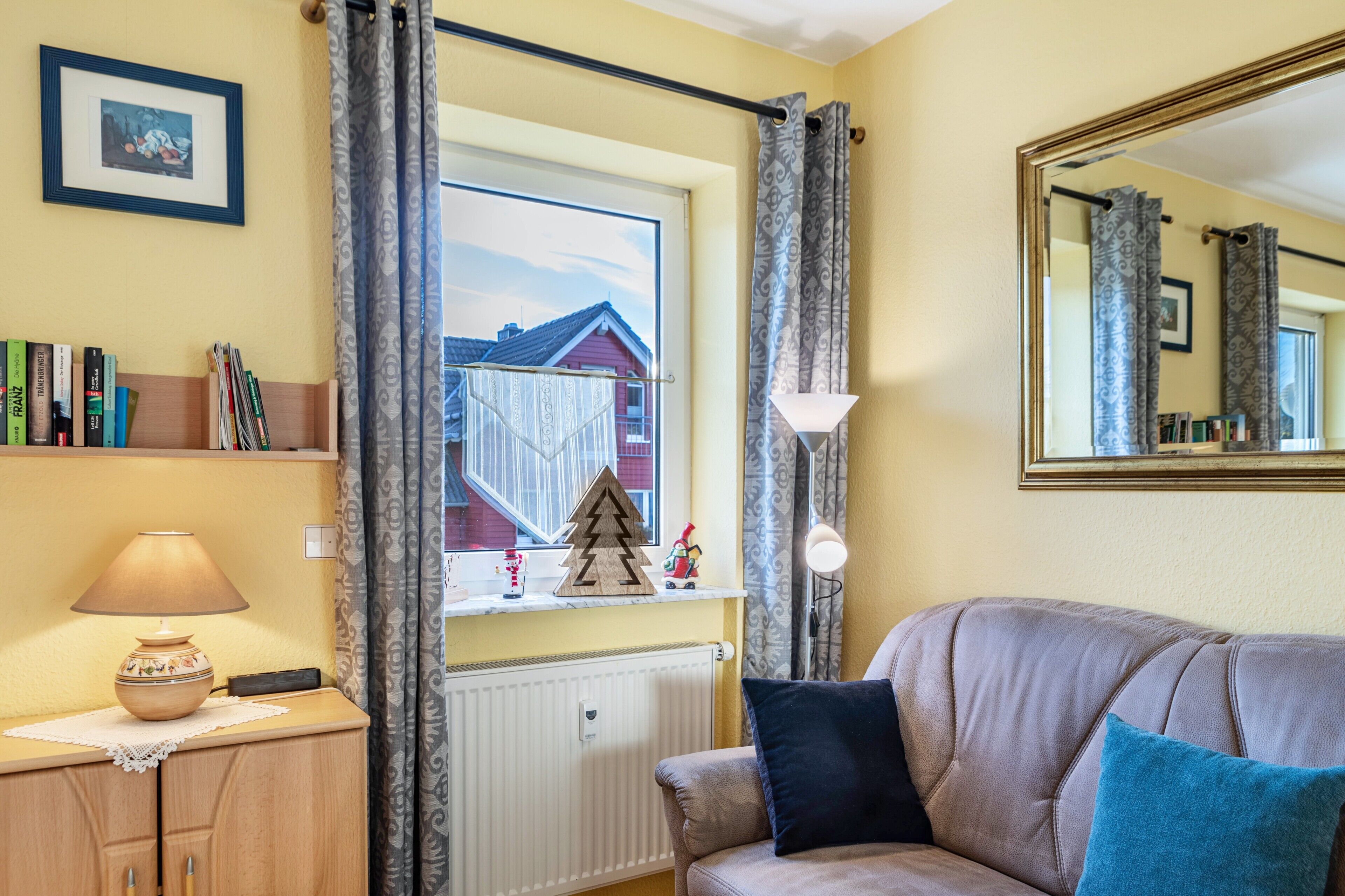 Un salon avec un canapé, une lampe, un radiateur, une fenêtre avec des rideaux, une étagère remplie de livres et un tableau encadré accroché au mur.