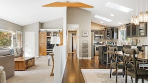 A spacious living area with a sofa, coffee table, and a wooden dining table with chairs. A kitchen with dark cabinets and a microwave is visible in the background. - Stunning Views + Arcade Games | Serene Retreat | Cabin in the Sky by AvantStay (Leicester)