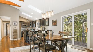 A spacious dining area with a wooden table and chairs, a kitchen in the background, and a large sliding glass door leading to a balcony with a view of trees. - Stunning Views + Arcade Games | Serene Retreat | Cabin in the Sky by AvantStay (Leicester)