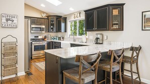 A modern kitchen with dark cabinets, a large island with bar stools, stainless steel appliances, and a window providing natural light. - Stunning Views + Arcade Games | Serene Retreat | Cabin in the Sky by AvantStay (Leicester)