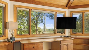 A wooden desk with a flat-screen TV, large windows with a view of trees, and a lamp. - Stunning Views + Arcade Games | Serene Retreat | Cabin in the Sky by AvantStay (Leicester)