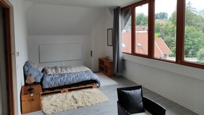Et soveværelse med en seng, et trænatbord, et lille bord, en stol og et stort vindue med udsigt til bygninger og træer.