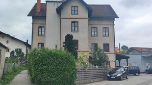 En toetagers bygning med en beige facade, flere vinduer og et overdækket parkeringsområde med biler.