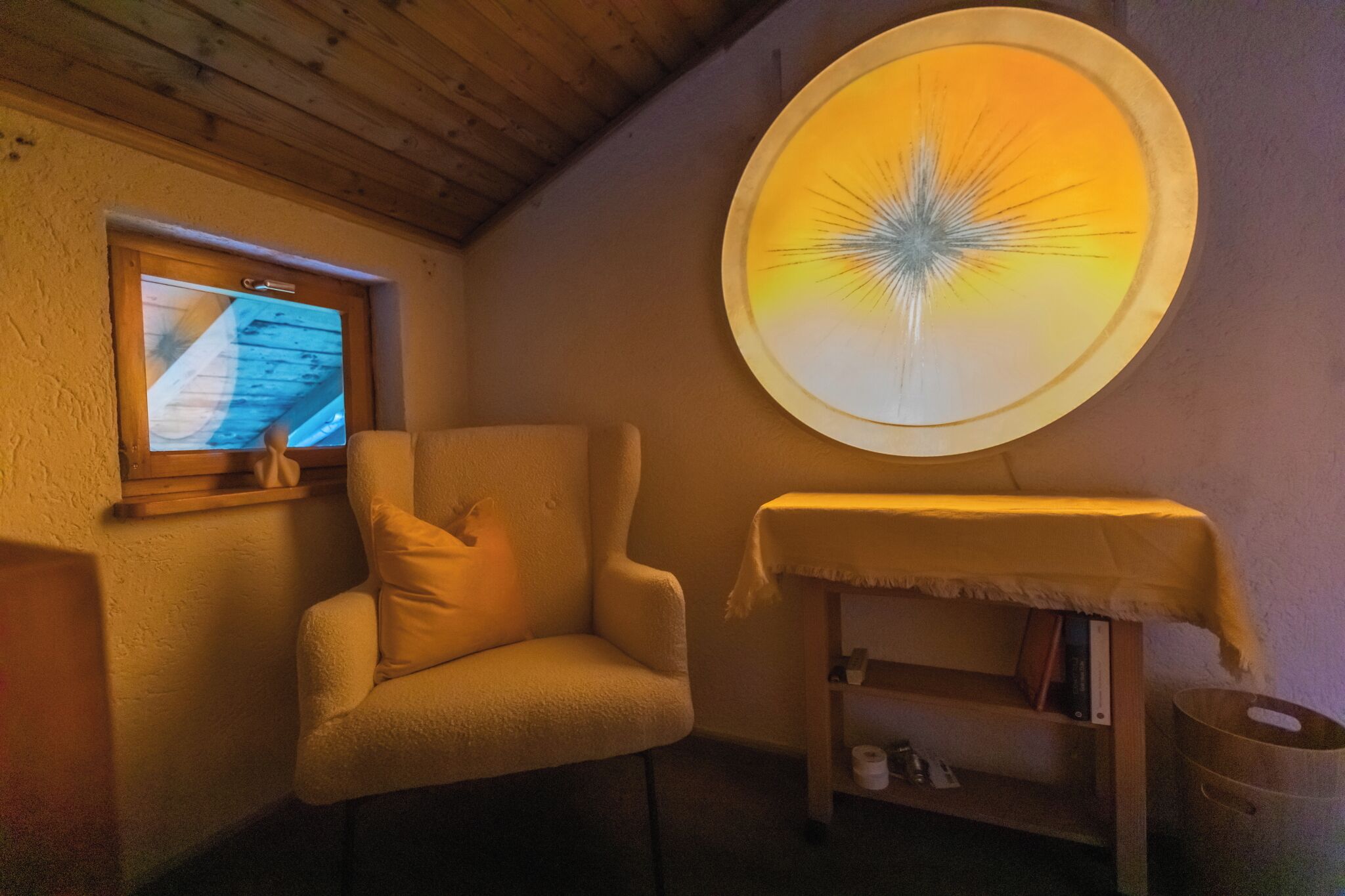 A cozy room with a plush armchair, a small table with a cloth, a wooden shelf with books, and a wall-mounted projector screen.