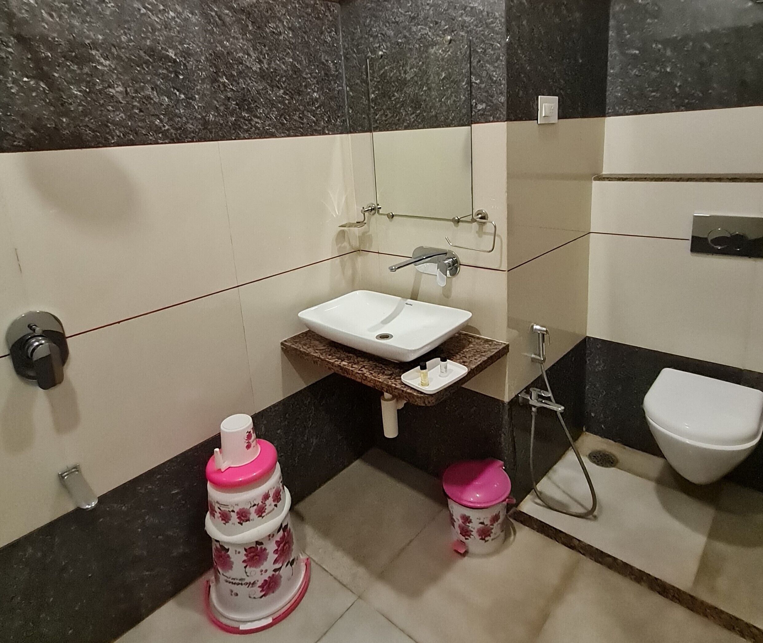 Een badkamer met een witte wastafel, een spiegel, een toilet en een douche. Er staan twee roze prullenbakken en een handdoekhouder.