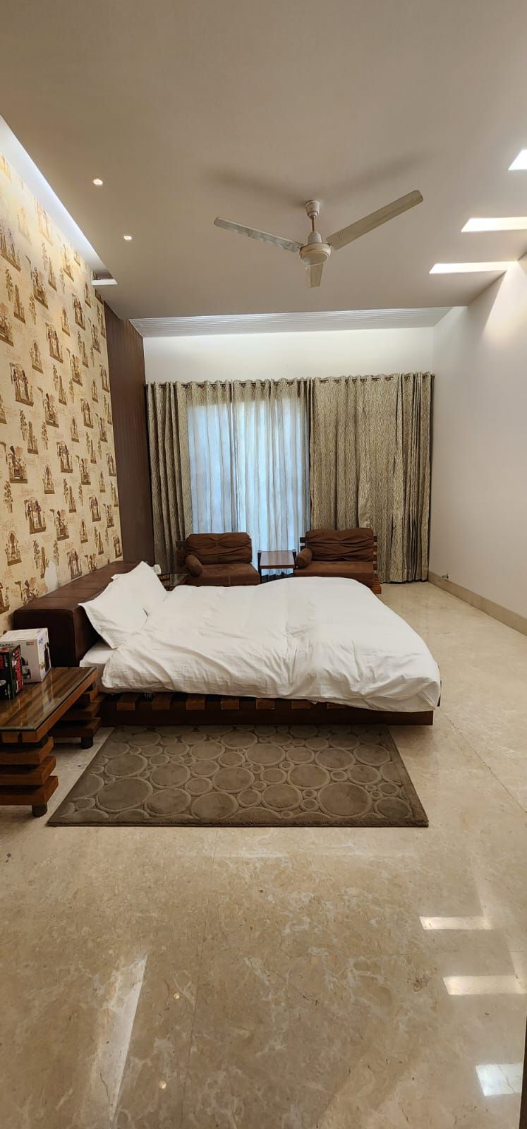 A bedroom with a large bed, a wooden nightstand, a ceiling fan, and a window with curtains.