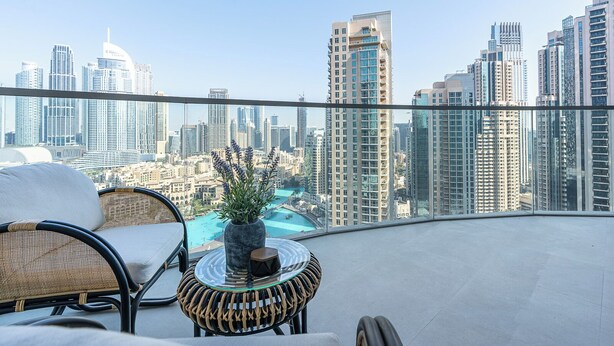 Un balcón con mobiliario de mimbre, una mesa de vidrio y vistas a un paisaje urbano con rascacielos y una piscina.