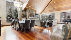 A spacious living area with a high ceiling, wooden beams, a stone fireplace, and large windows offering scenic mountain views. - Cascade Village - Westhaven Condo Sleeps 24 (Vail)