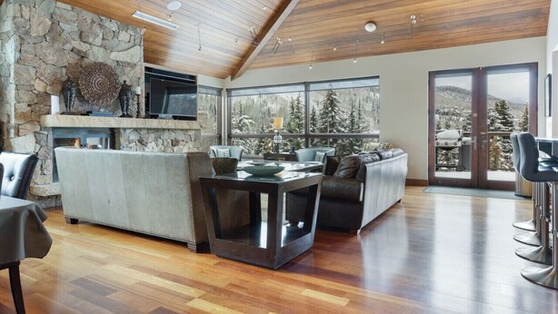 A spacious living area with a stone fireplace, large windows offering a view of snow-covered trees, and a high wooden ceiling. - Cascade Village - Westhaven Condo Sleeps 24 (Vail)
