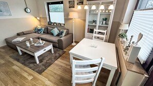 Ein modernes Wohnzimmer mit einer Couch, einem Couchtisch und einem Esstisch. Ein Bücherregal und ein Fenster mit Jalousien sind ebenfalls vorhanden.