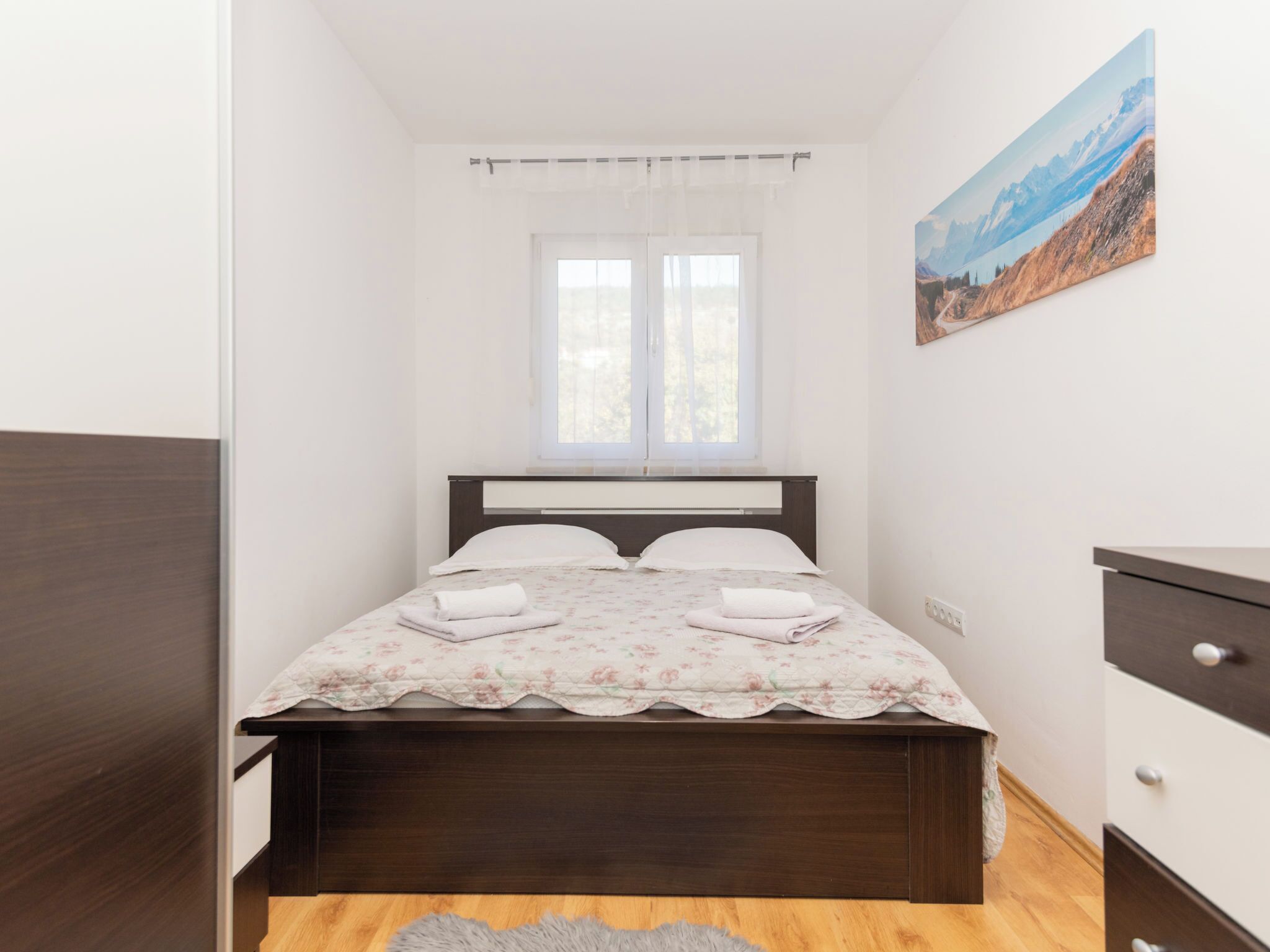 A bedroom with a wooden bed, white curtains, a painting of a mountain landscape, and a wooden dresser.