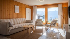 A cozy living room with a wooden wall, a sofa, two armchairs, a coffee table, and a view of mountains through large windows. - Seya 15 by Interhome (Ovronnaz)