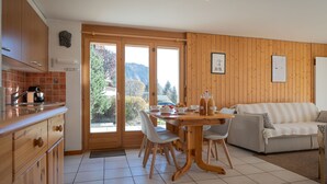 A modern kitchen with wooden cabinets, a dining table with chairs, a sofa, and a view of a mountainous landscape through large glass doors. - Seya 15 by Interhome (Ovronnaz)