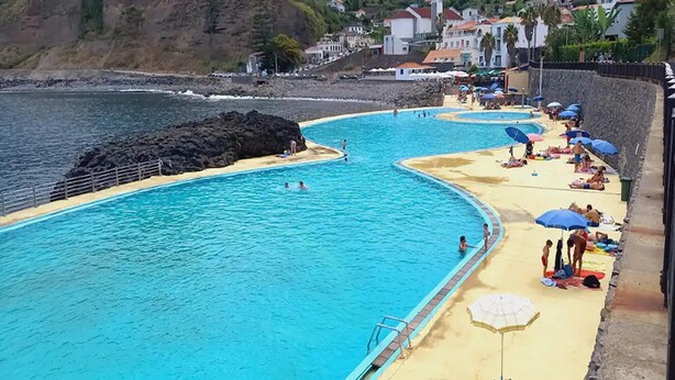 Una piscina a la costa amb aigua cristal·lina blava, envoltada per una platja de sorra i una paret de pedra.