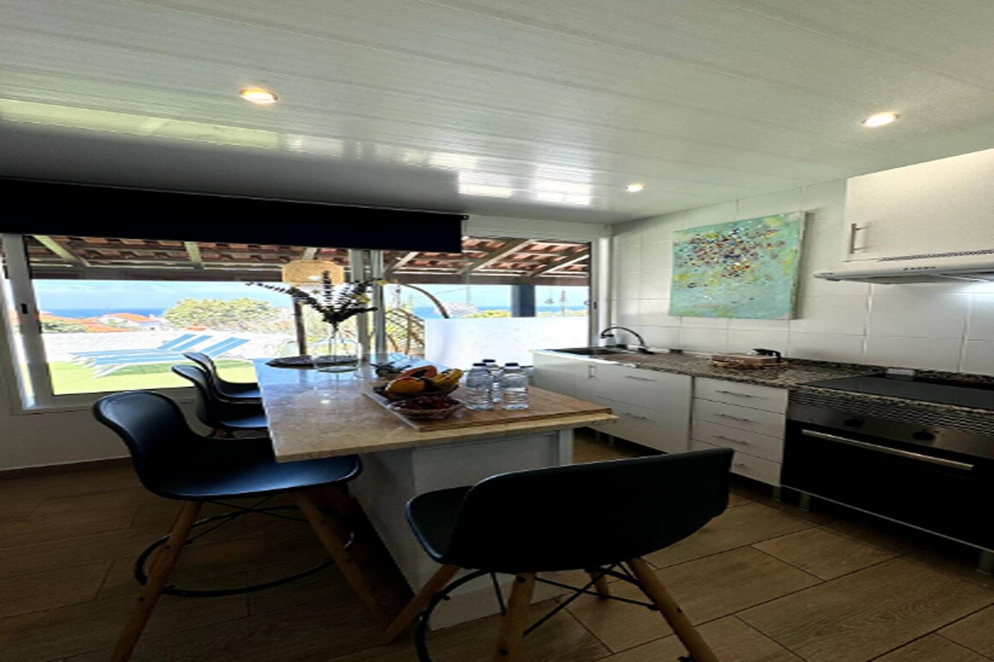 A modern kitchen with a large island, bar stools, a view of the outdoors, and a painting on the wall.