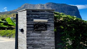 Una costruzione in pietra con una targa e un piccolo cartello, immersa nel verde con montagne sullo sfondo.