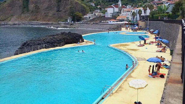Una piscina vista mare con spiaggia sabbiosa e ombrelloni, circondata da case e scogliere.