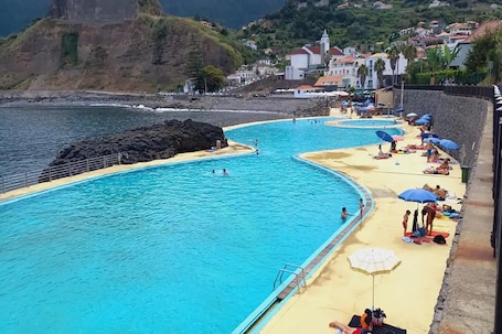 A coastal swimming pool with a sandy beach and umbrellas, surrounded by houses and cliffs.