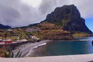 A coastal area with a prominent rocky hill, a beach with a rocky shoreline, and several buildings with red roofs. - Studio in Porto da Cruz with Ocean Views (Porto da cruz , Machico)