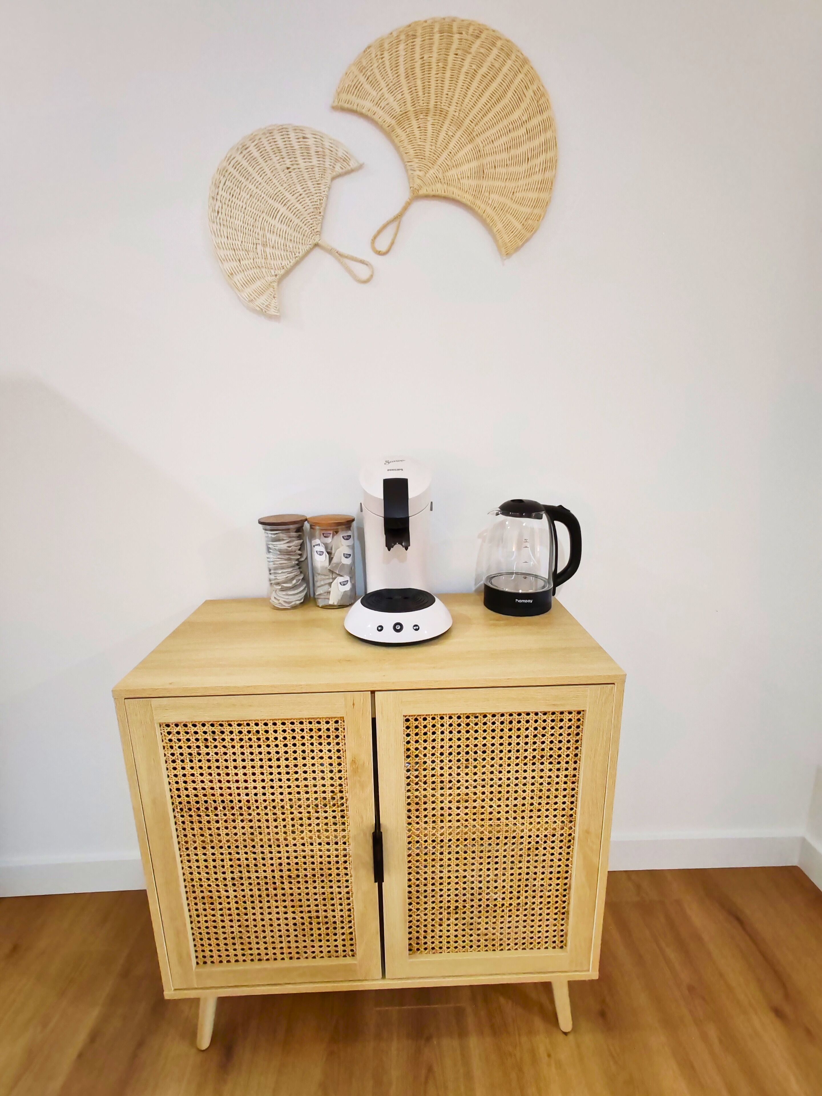 Eine Holzkabine mit zwei Türen, eine weiße Kaffeemaschine, ein schwarzer Wasserkocher und zwei gewebte Wandbilder.