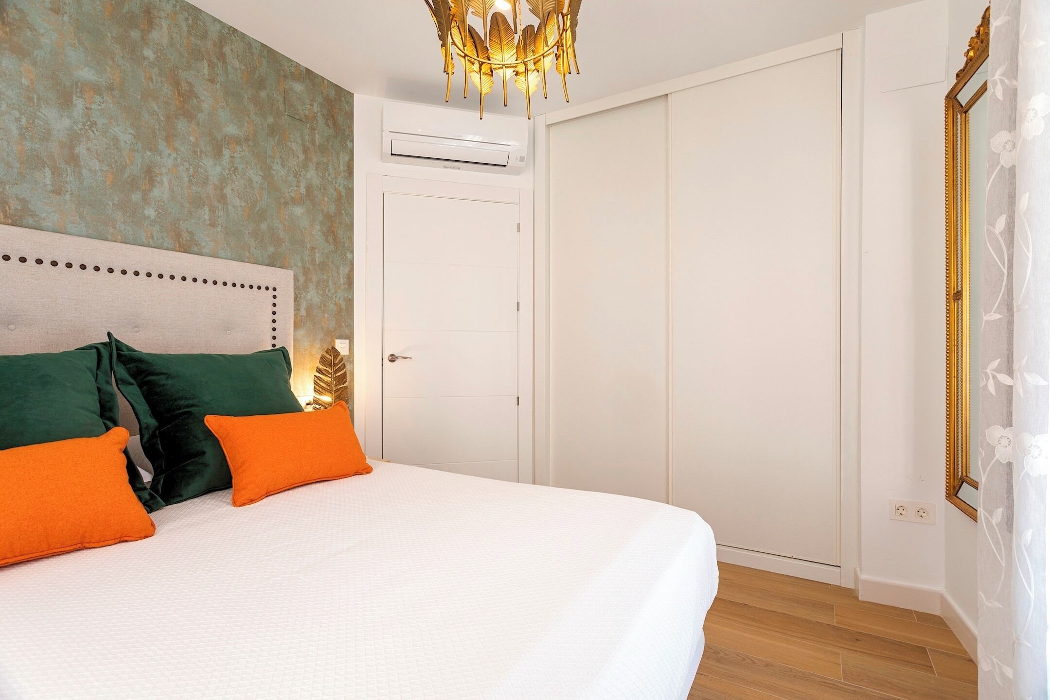 Una cama bien tendida con ropa de cama blanca y verde, un aire acondicionado de pared y un amplio armario con espejo.