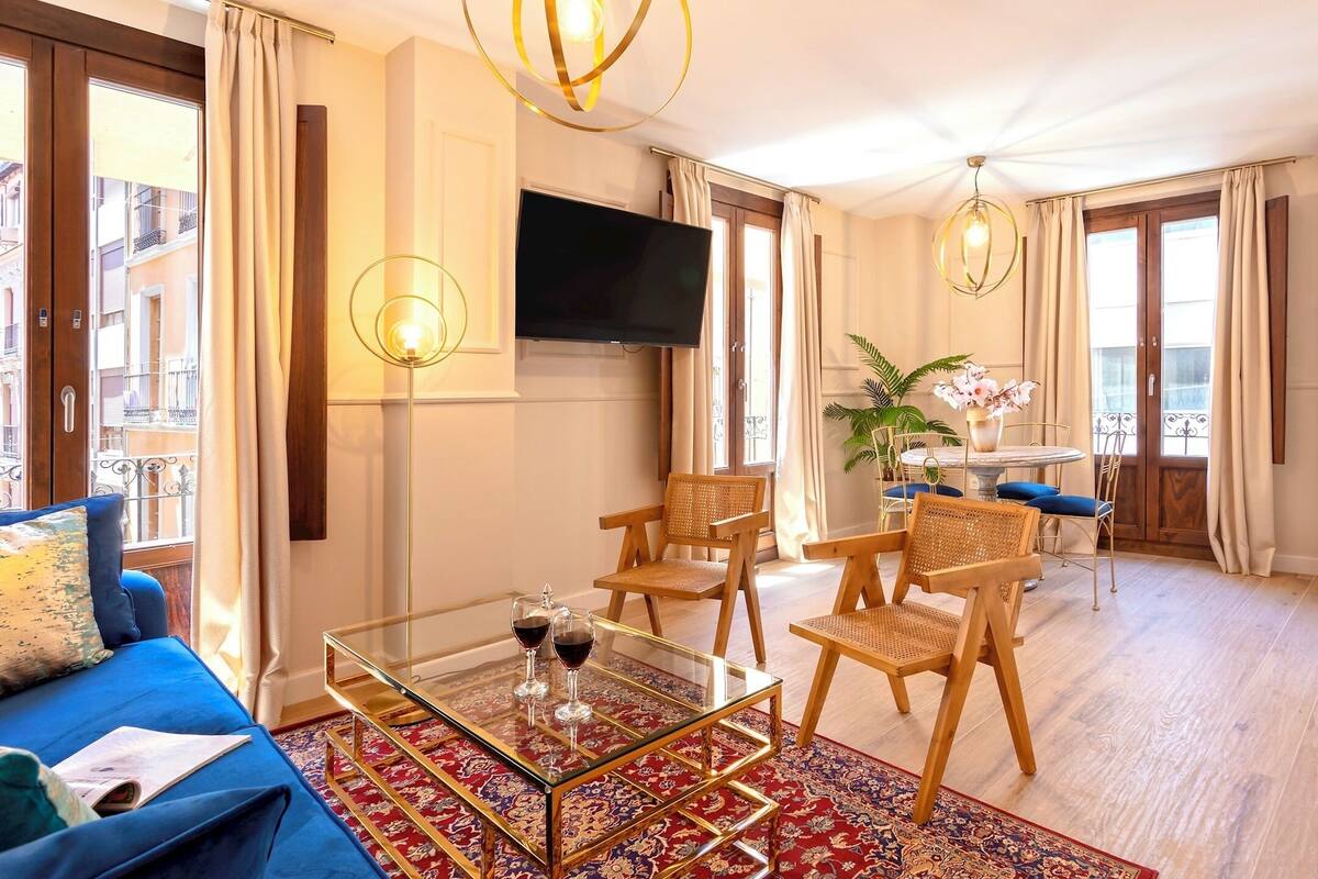 A hotel room with a blue sofa, a glass coffee table with wine glasses, a TV mounted on the wall, and a dining area with wicker chairs and a table.