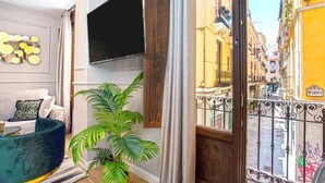 Ein modernes Wohnzimmer mit großem Fernseher, einem grünen Sofa, einer Topfpflanze und Blick auf eine enge Straße durch einen Balkon mit schmiedeeisernem Geländer.