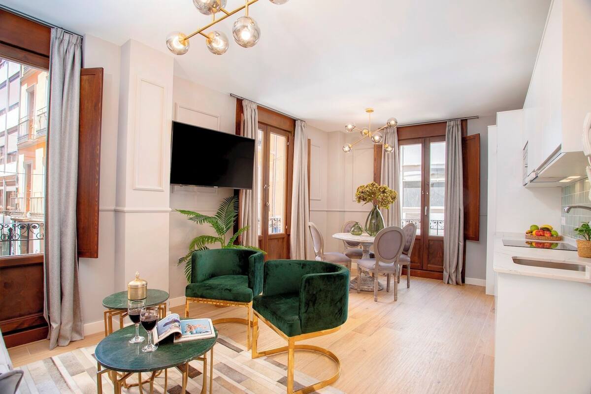 A modern living room with a green sofa, a coffee table, and a TV mounted on the wall. There is a dining area with a table and chairs, and a kitchen area with a sink and countertop.