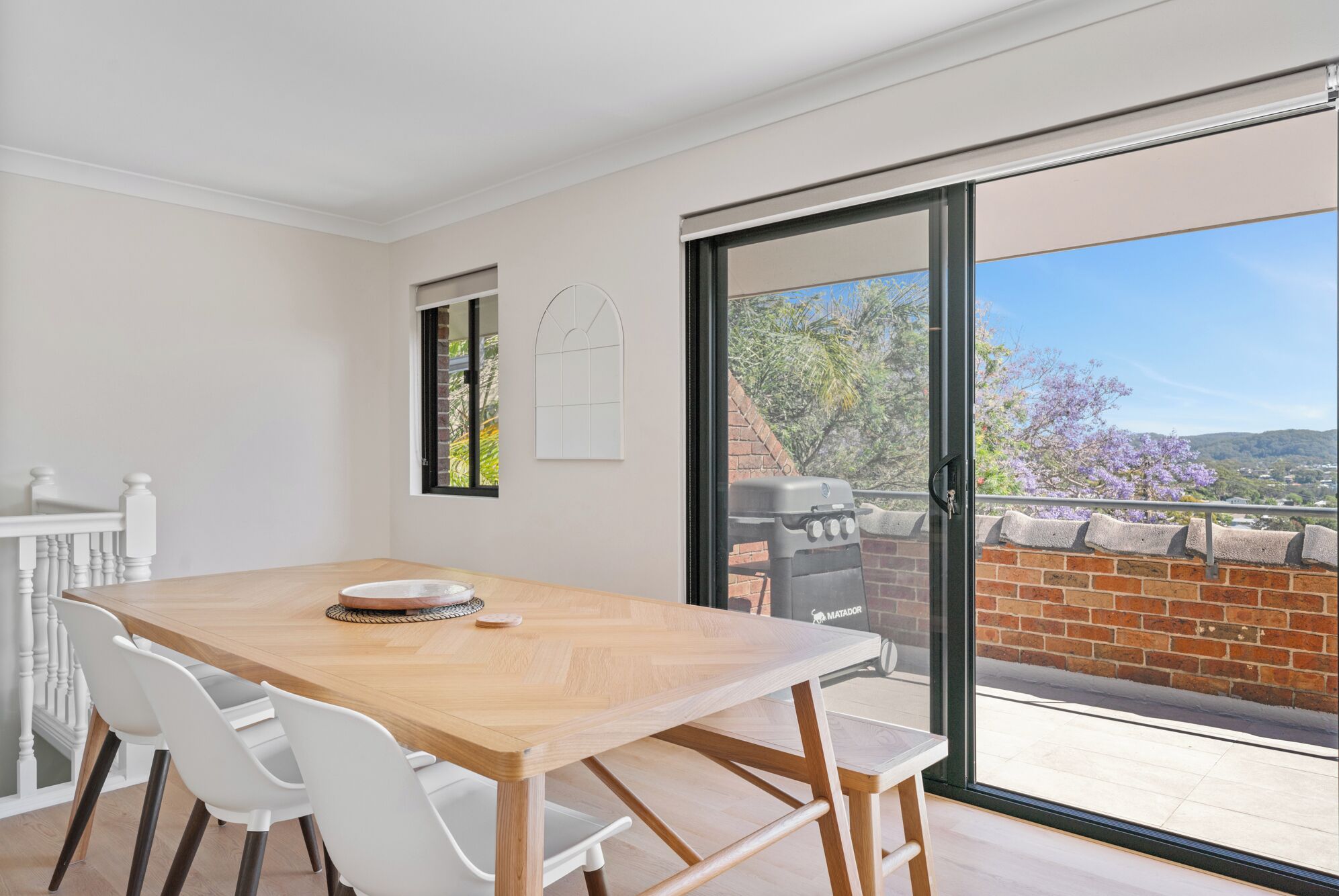 A dining area with a wooden table and white chairs, a sliding glass door leading to a patio with a barbecue grill.