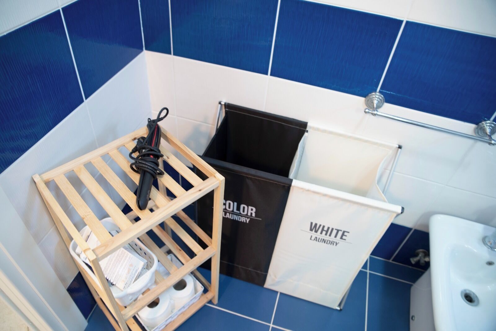 A bathroom with blue tiles, a wooden shelf with toiletries, and two laundry bags labeled "COLOR LAUNDRY" and "WHITE LAUNDRY".