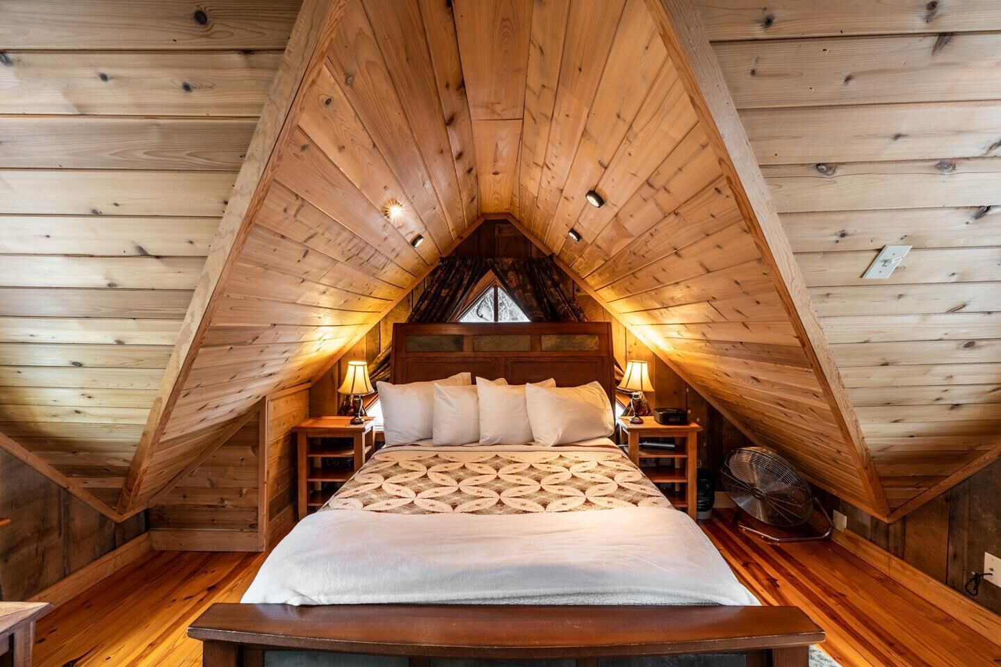 Une chambre confortable située sous les toits, avec un plafond en pente en bois, un grand lit recouvert d’une couvre-lit à motifs, deux lampes de chevet et une petite table.