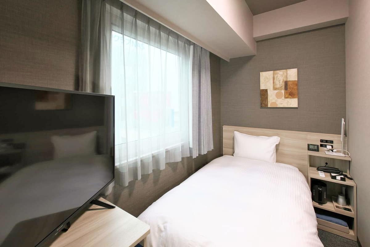 A modern hotel room with a bed, a TV, a bedside table, a nightstand, and a window with sheer curtains.