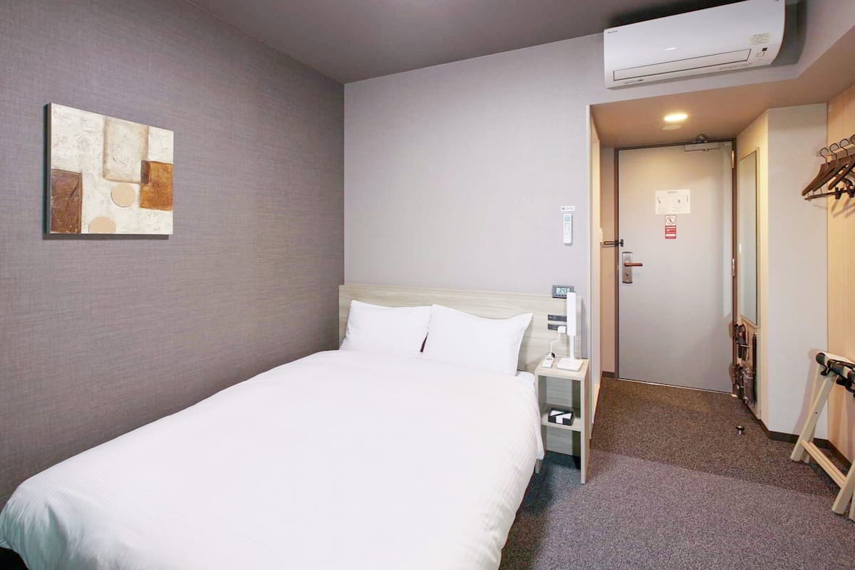A hotel room with a bed, a bedside table, an air conditioning unit, a painting on the wall, and a door leading to a hallway.