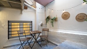 En liten sittplats utomhus med ett bord och stolar, ett fönster med persienner och dekorativa, flätade korgar på väggen.