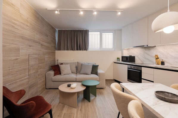 A modern living room with a sofa, coffee table, and chairs, a kitchen with a stove and cabinets, and a window with curtains.