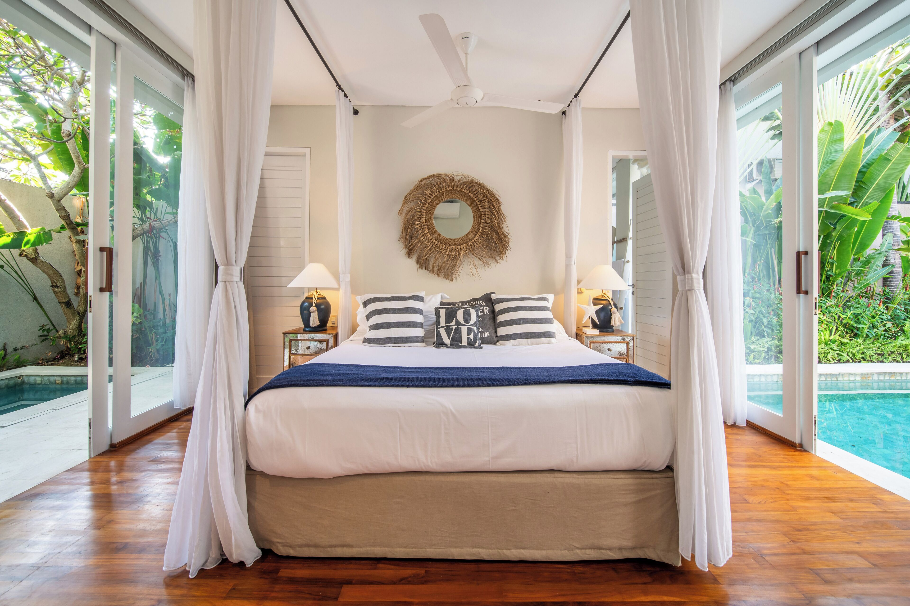 A bedroom with a bed, curtains, a ceiling fan, a bedside table with a lamp, and a view of a swimming pool.