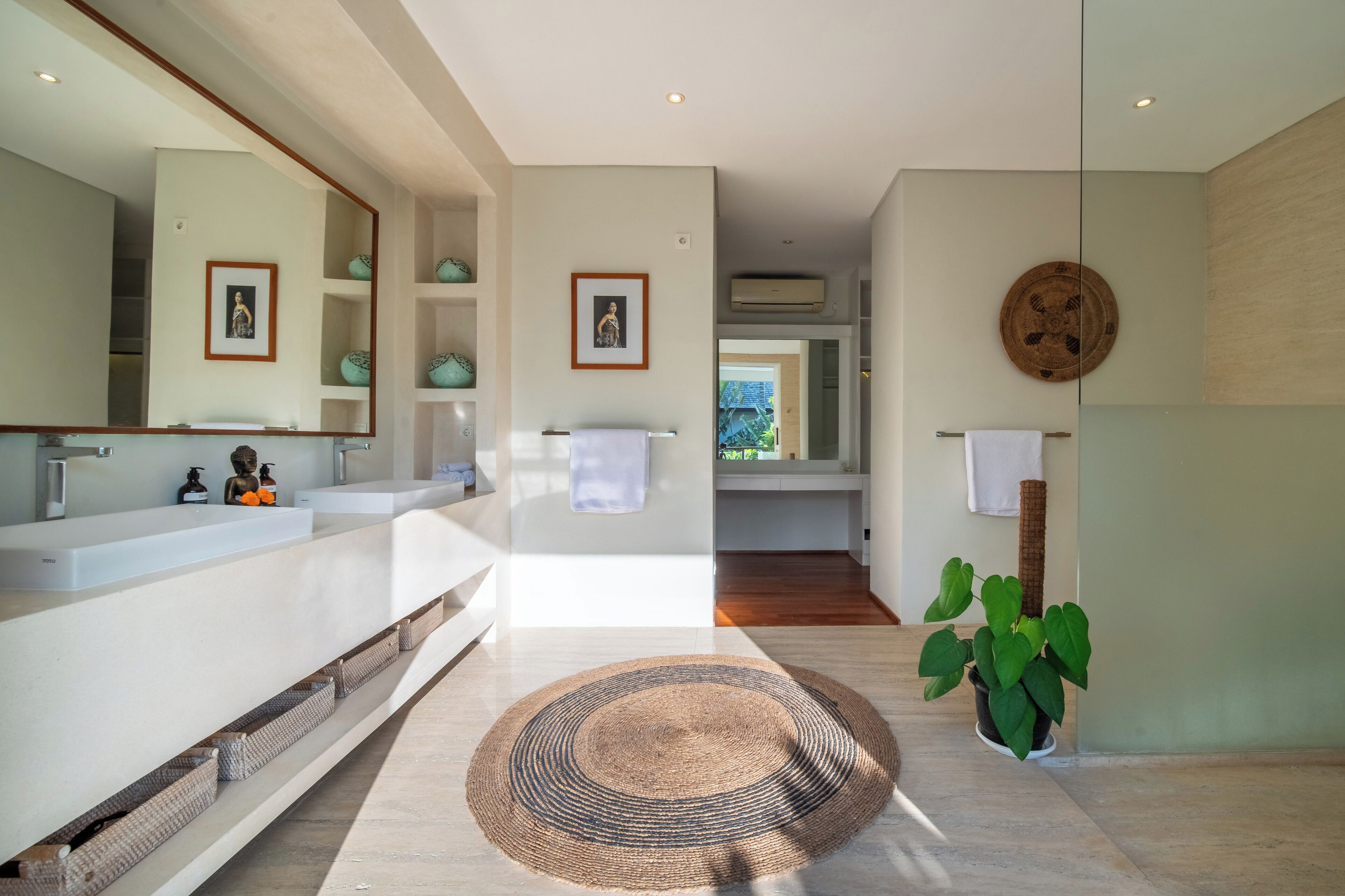 A modern bathroom with a large mirror, two sinks, a round woven rug, and a potted plant.
