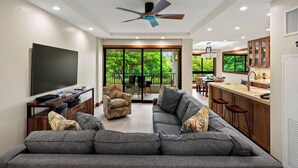 A modern living room with a grey sofa, a flat-screen TV, a ceiling fan, and a large window offering a view of greenery.