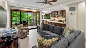 A modern living room with a grey sofa, a coffee table, and a bookshelf. There is a balcony with a view of greenery.