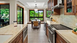 A modern kitchen with a central island, wooden cabinets, and large windows offering a view of greenery.