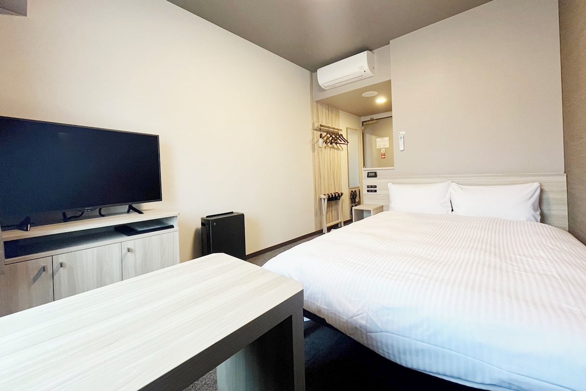 A hotel room with a bed, a TV, a desk, a wardrobe, and an air conditioning unit.