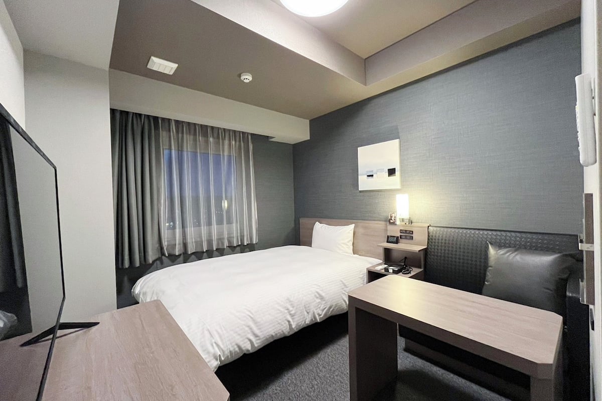A hotel room with a bed, bedside table, lamp, and a window with curtains.