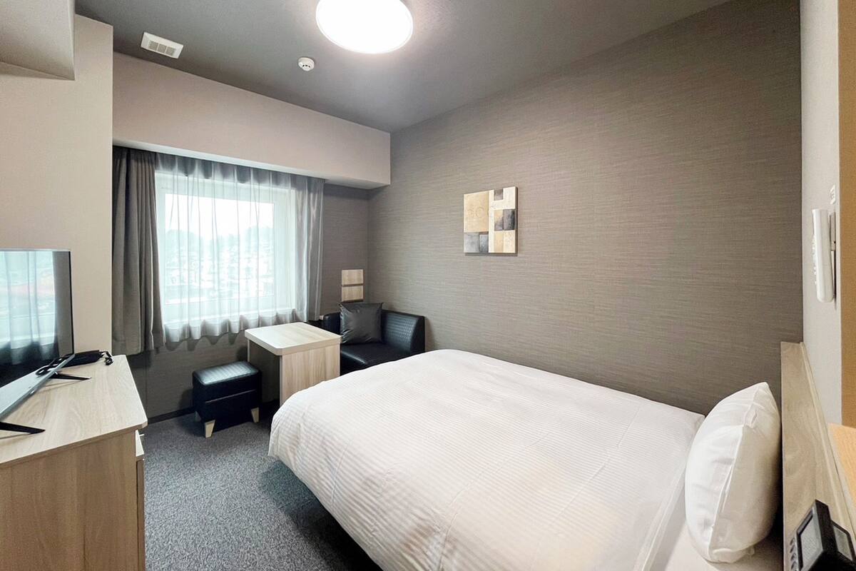 A hotel room with a bed, a desk, a chair, a TV, and a window with curtains.