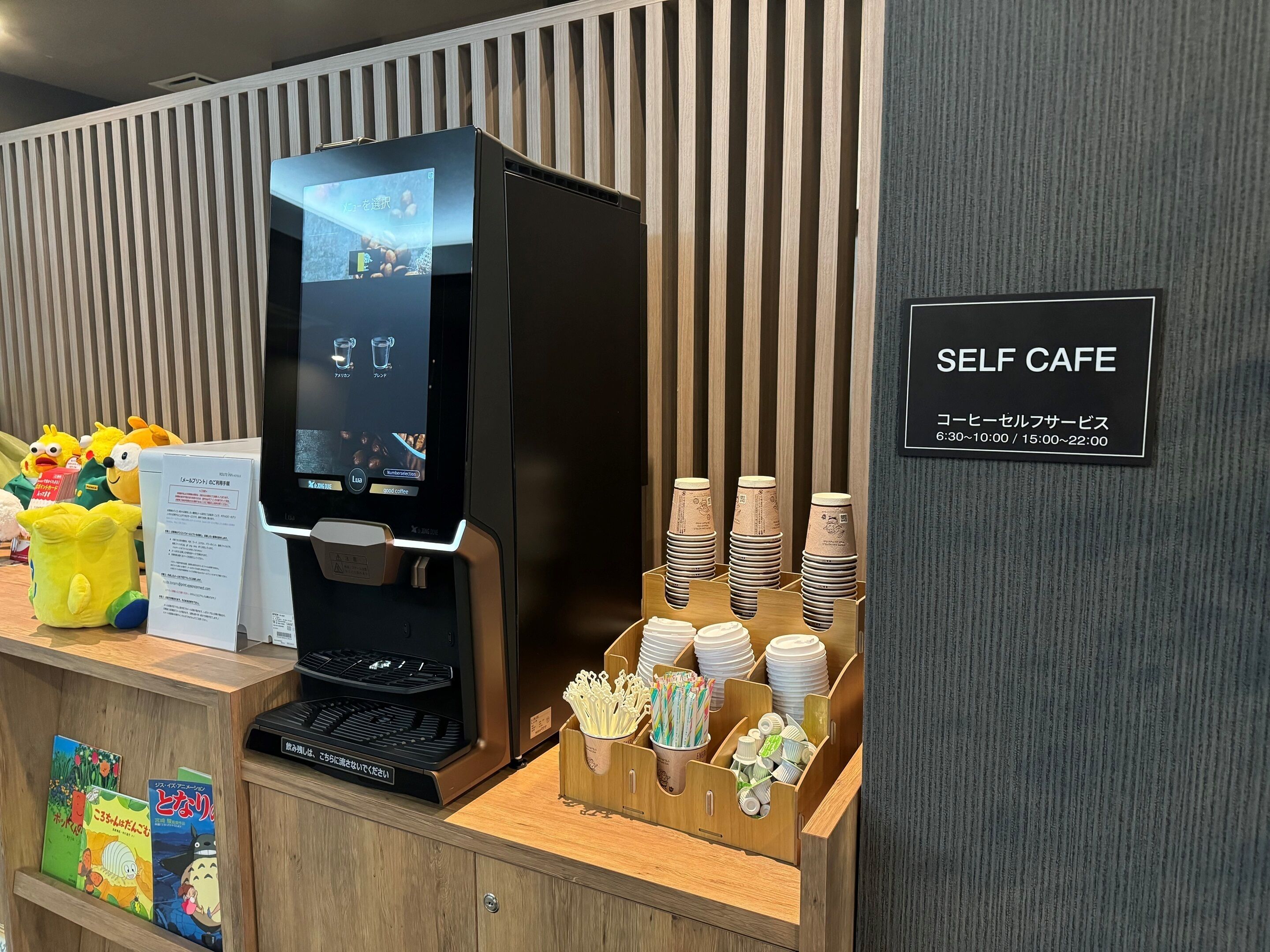 Una estació de cafè de servei automàtic amb un panell digital de menú, gots d'un sol ús i un cartell que indica l'horari d'obertura.