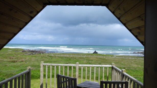 Een houten terras met tafel en stoelen, met uitzicht op een grasveld en de oceaan.